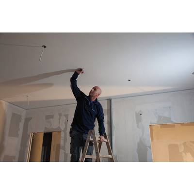 A man is standing on a ladder and working on the ceiling of an unfinished room. Walls are partially plastered.