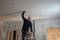 A man is standing on a ladder and working on the ceiling of an unfinished room. Walls are partially plastered.