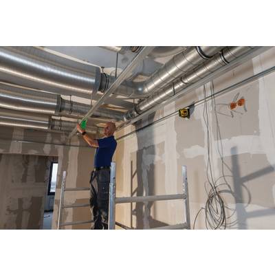 A worker is standing on scaffolding and installing ventilation pipes on the ceiling of an unfinished building area.