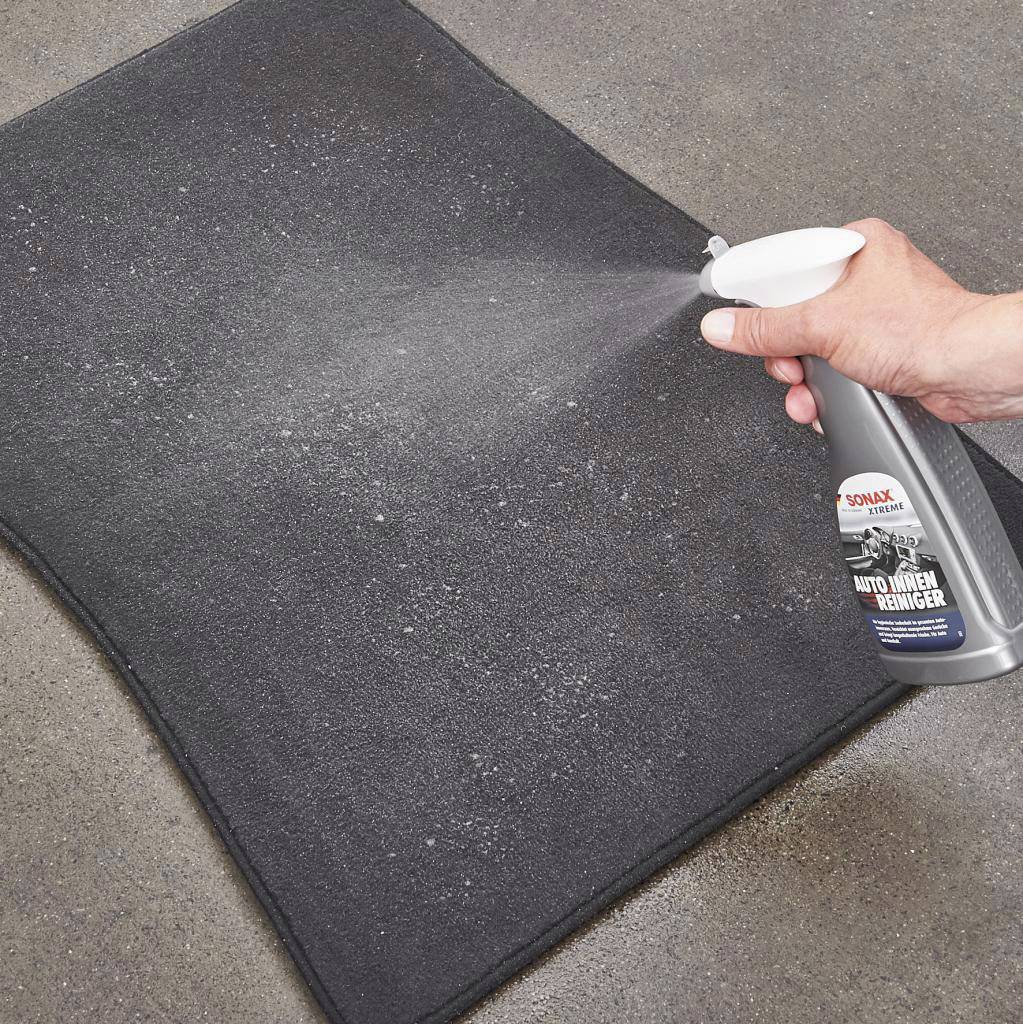 A hand sprays car interior cleaner from a bottle onto a black floor mat to remove dirt.