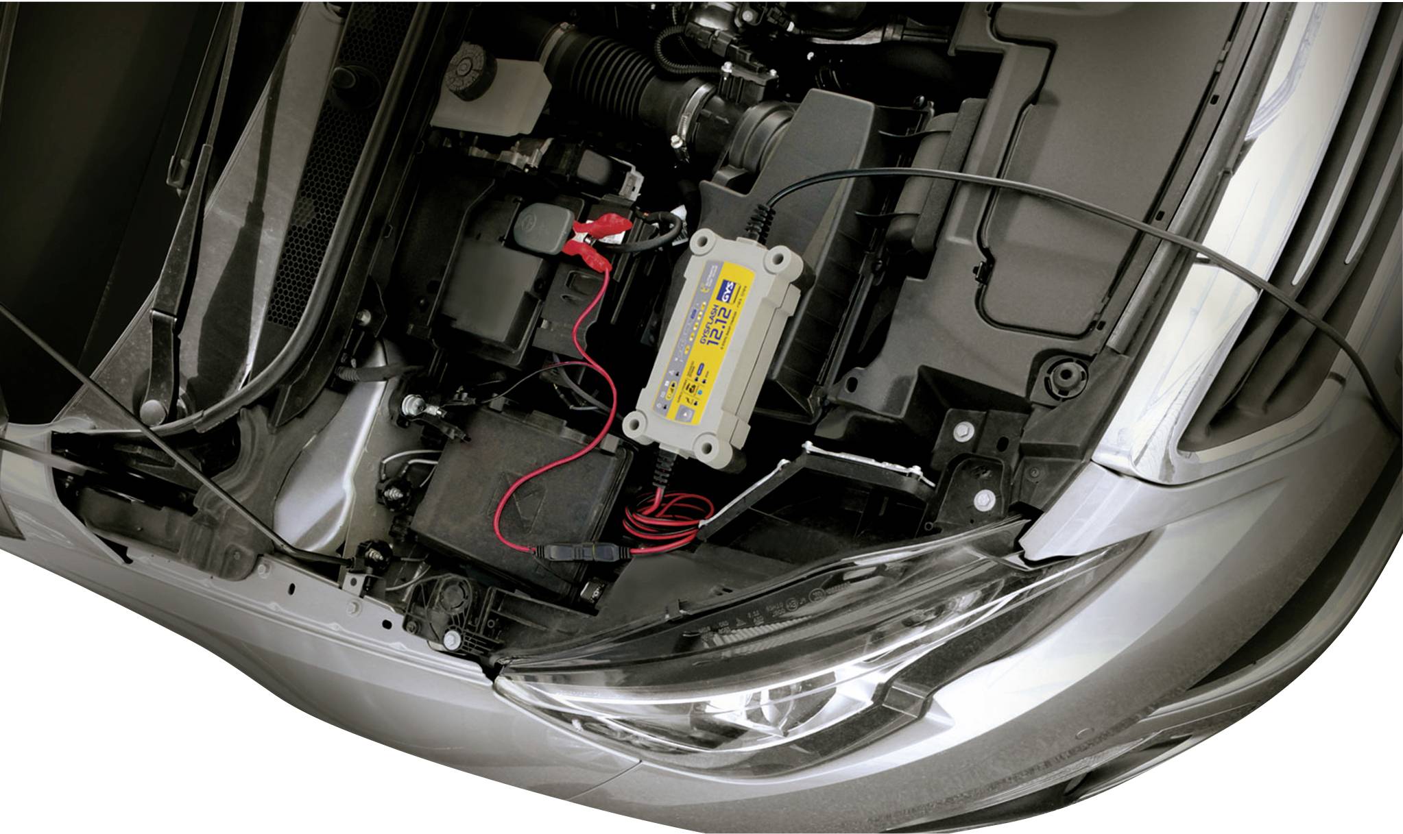 A car charger is charging a car battery in the open engine compartment of a vehicle. Red and black cables are connected.