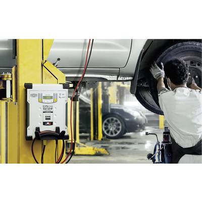 A mechanic inspects a raised car in a workshop environment. A battery charger is visible in the foreground.