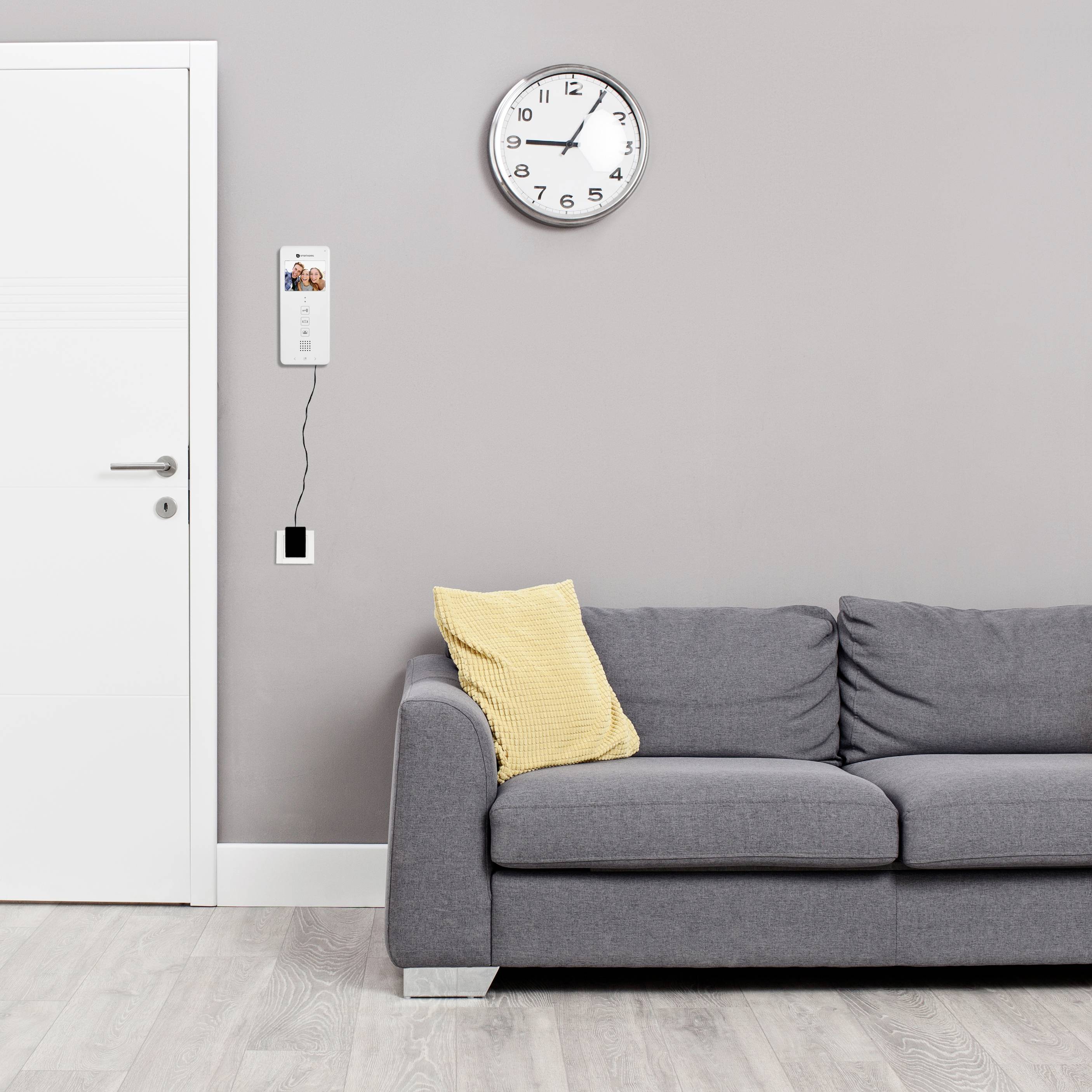 A grey sofa with a yellow cushion sits in a living room. A clock hangs on the grey wall, with an intercom beside it.