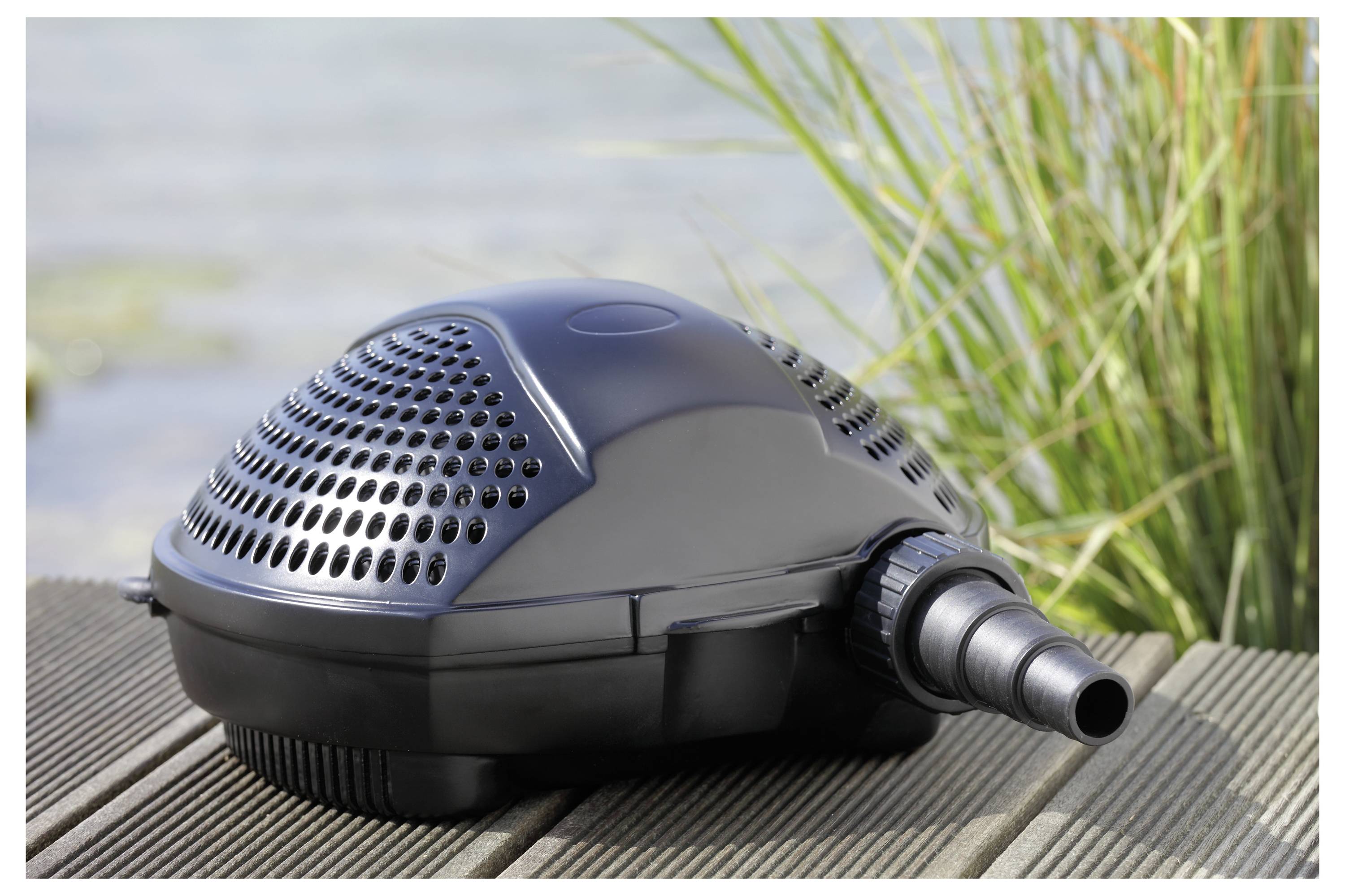 A black pond pump placed on wooden decking near a pond with tall grass in the background.