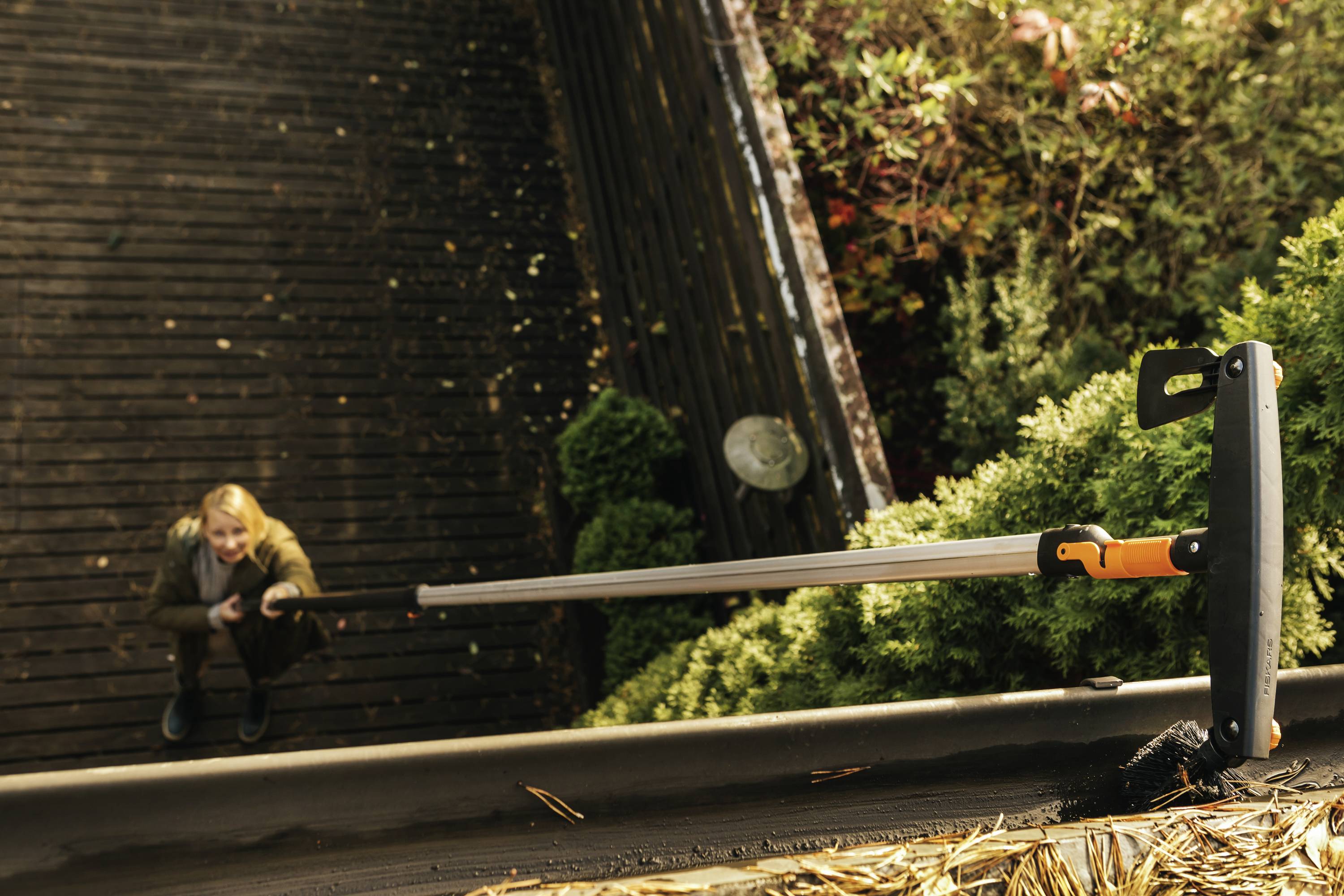 A person is standing below and looking up at a roof. A cleaning tool is being used to remove leaves from the guttering.