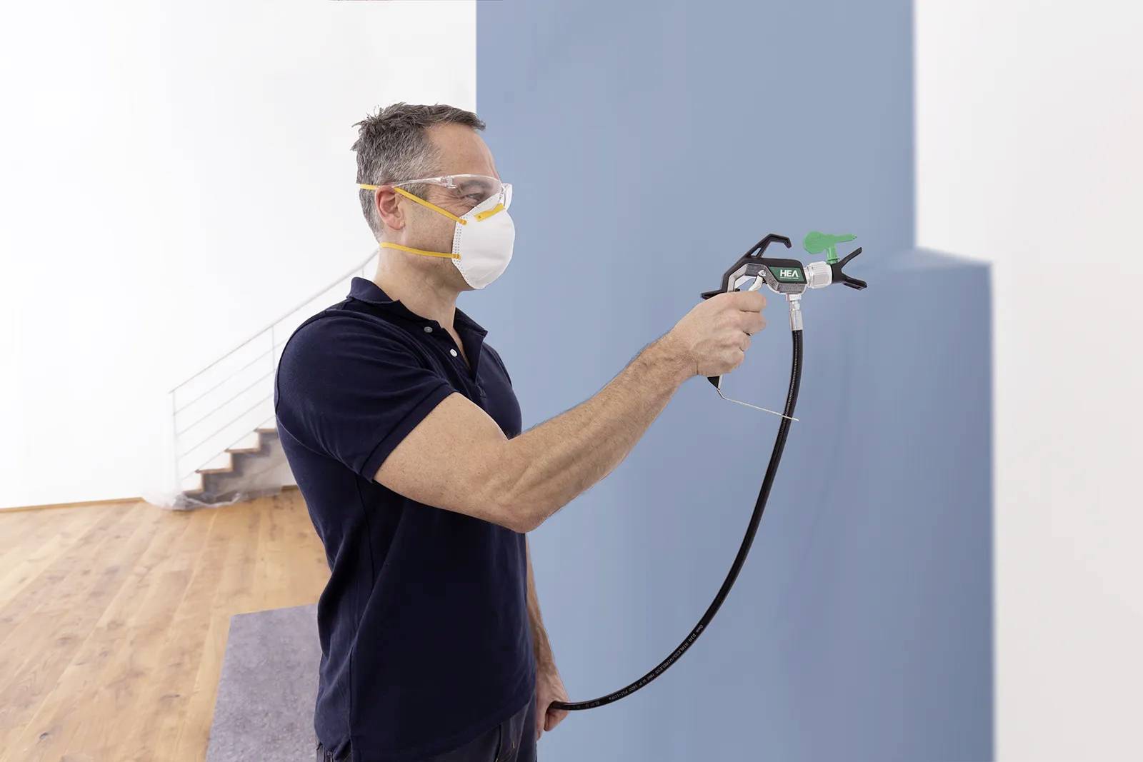 A man wearing safety goggles and a mask is spraying blue paint onto a wall in a room with a wooden floor and a staircase in the background.