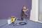 A man is kneeling on a carpet and spraying a wall purple with a spray gun. He is wearing a respiratory mask.