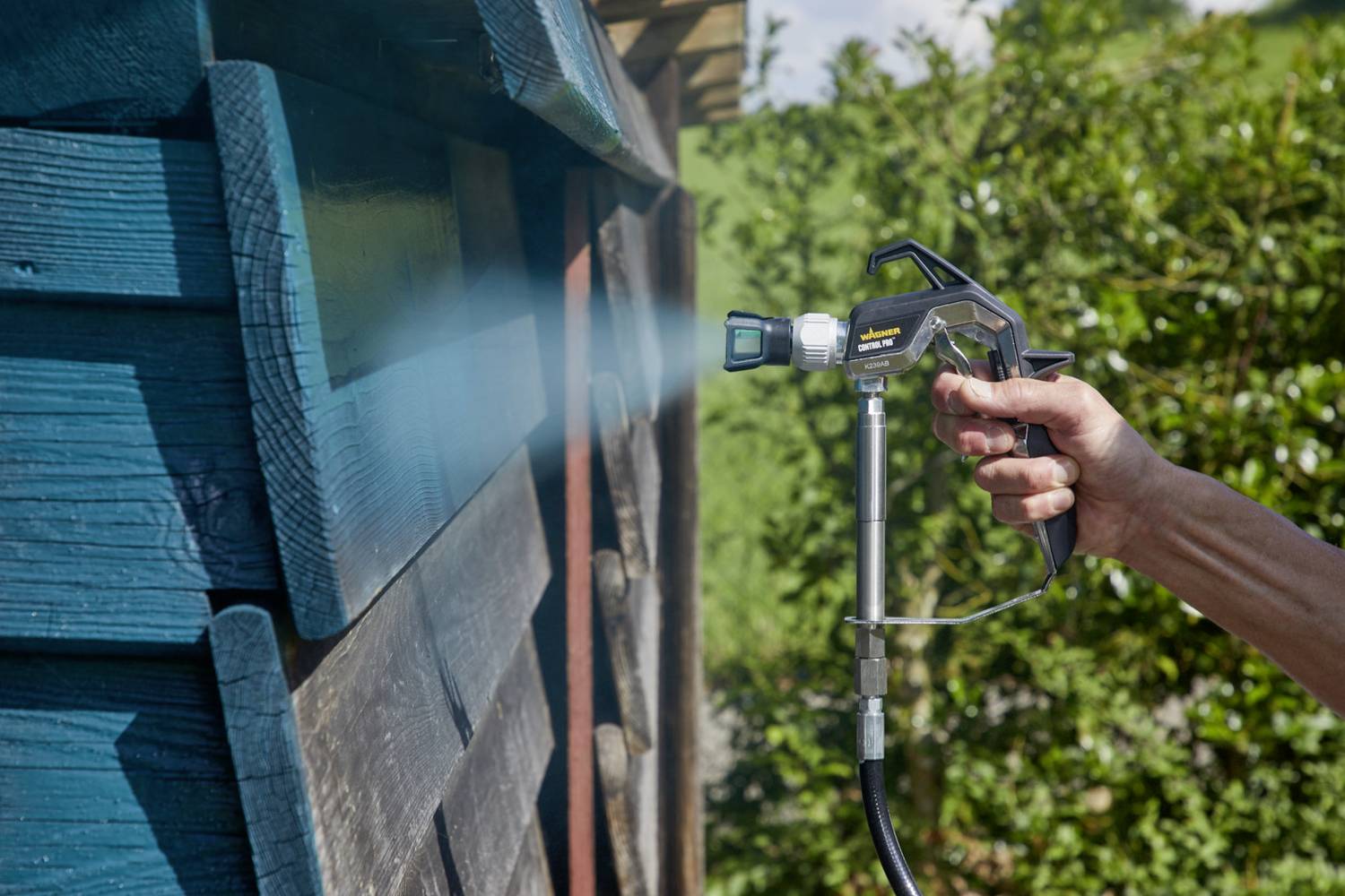 A man is painting a wooden house with blue paint outdoors. Next to him stands a yellow paint sprayer on the grass.