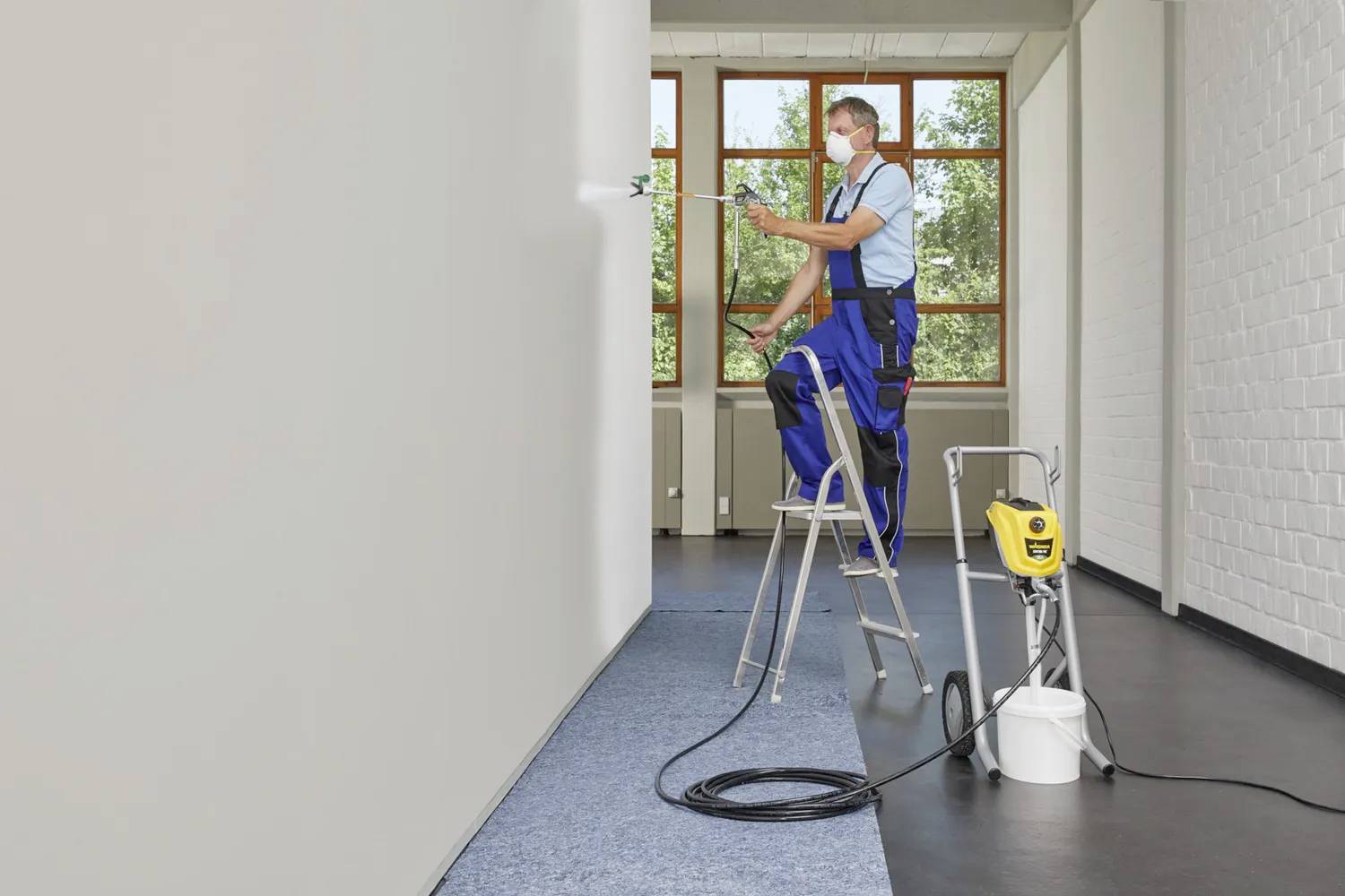 A tradesman wearing a respiratory mask is standing on a ladder and spraying white paint onto an interior wall in a bright room.