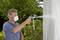 A man wearing a respiratory mask is spraying a light surface outdoors with a spray gun, surrounded by green vegetation.