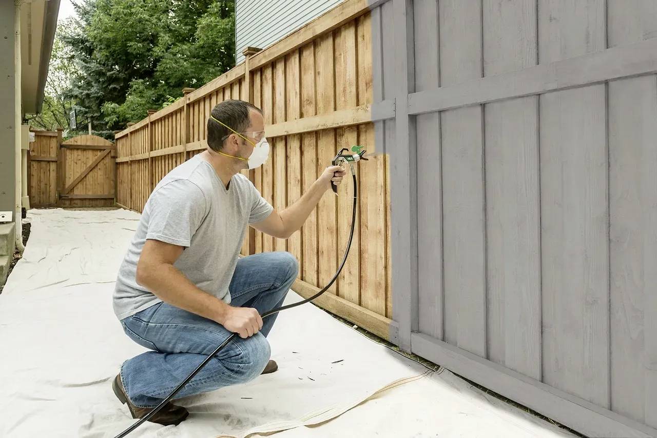 A person wearing a respiratory mask is spraying paint onto a wooden fence in the garden. It is a sunny, bright environment with trees in the background.