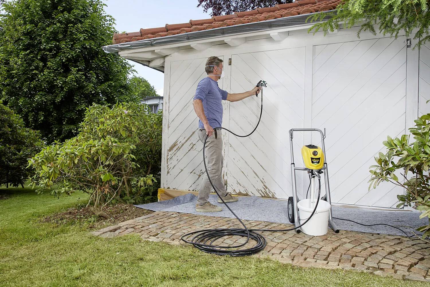 A man is painting a garage wall white with a spray device. The garage has a sloping roof and is surrounded by bushes.