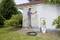 A man is painting a garage wall white with a spray device. The garage has a sloping roof and is surrounded by bushes.