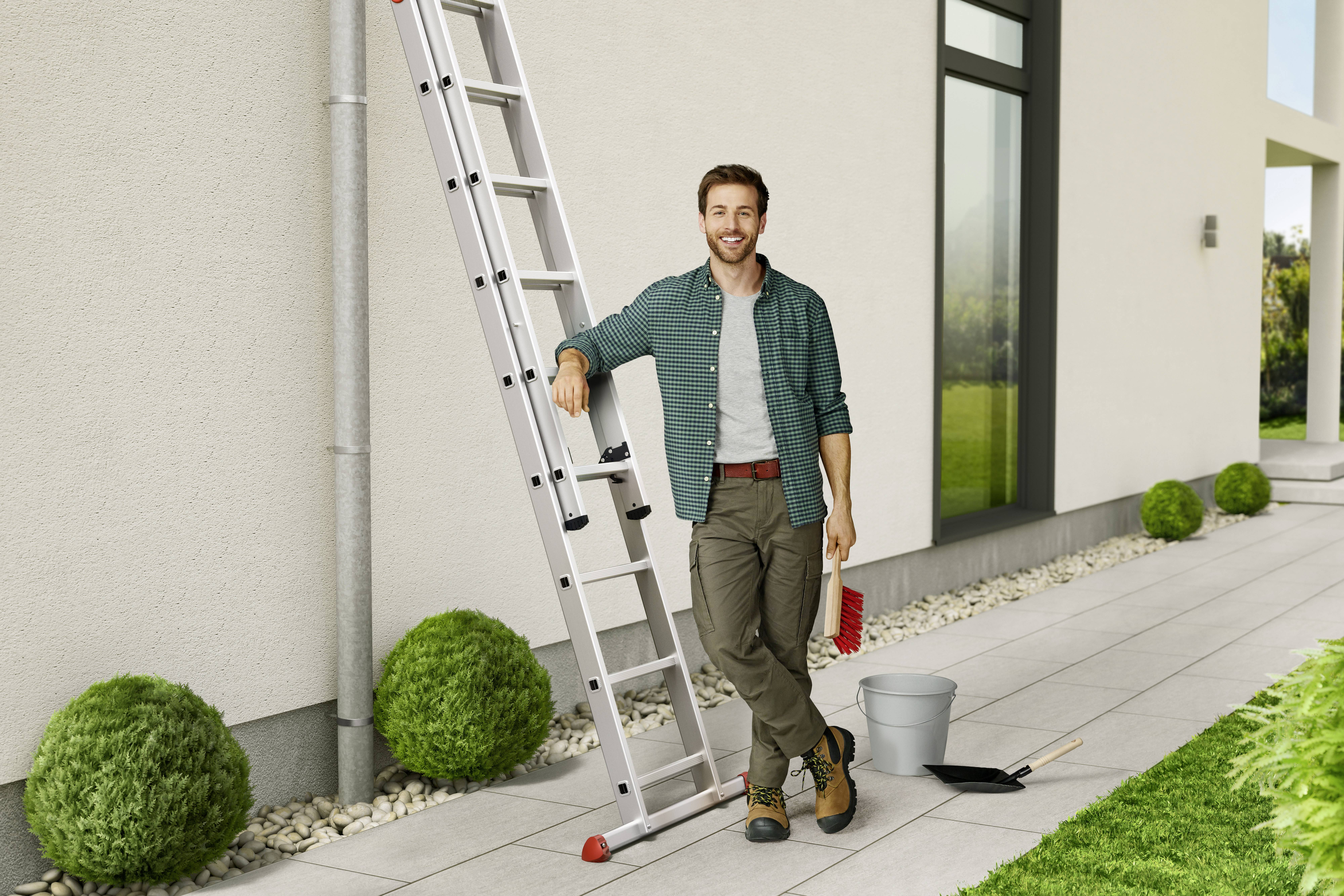 A man is standing smiling next to a ladder against a house wall, holding a red brush. A bucket and a shovel are beside him.
