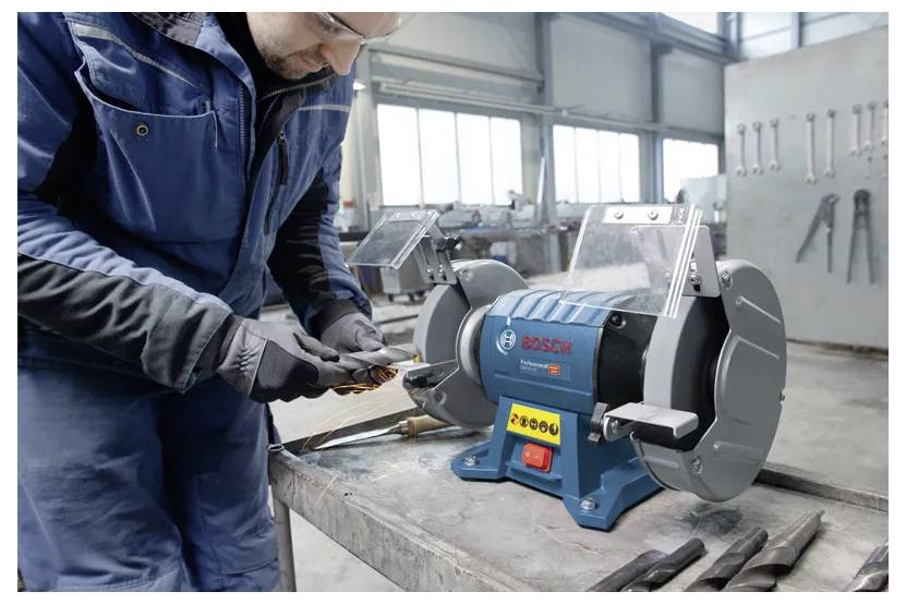 A person in protective gear sharpens a tool using a Bosch bench grinder in an industrial workshop setting.