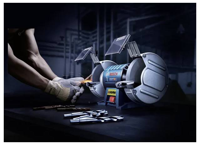 A person operates a Bosch bench grinder, sharpening a tool on the grinding wheel. Nearby, metal rods are arranged on a table.