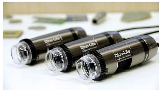 Three Dino-Lite digital microscopes are aligned on a table, showcasing their design. Blurred items are visible in the background.