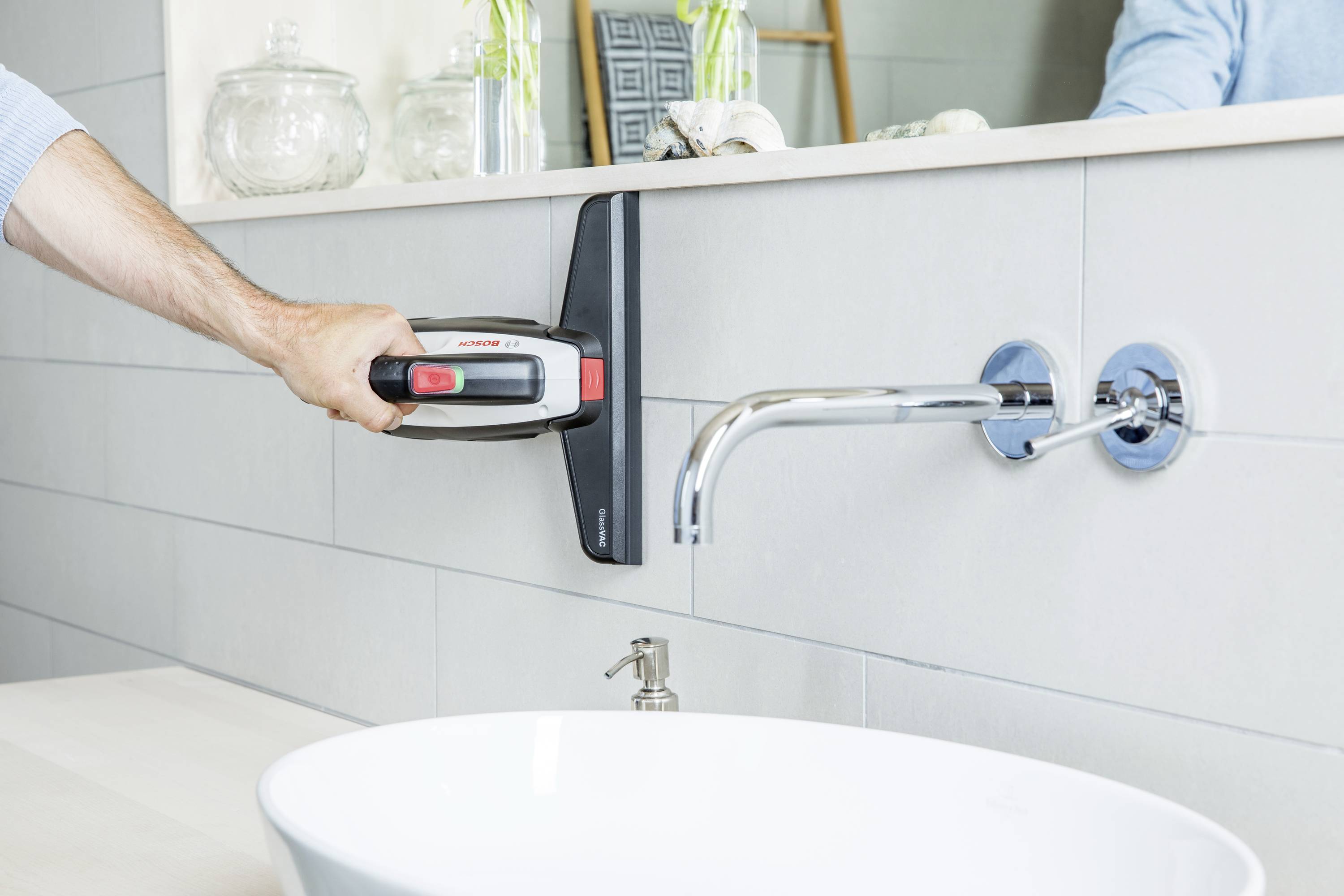 A person is cleaning a smooth, tiled bathroom wall with a handheld vacuum cleaner near a tap.