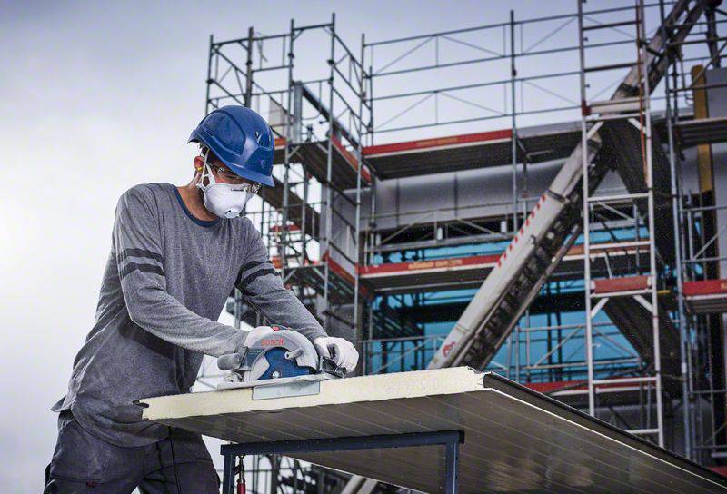 A man wearing a hard hat and mask is cutting a metal sheet with a saw, in front of scaffolding on a construction site.