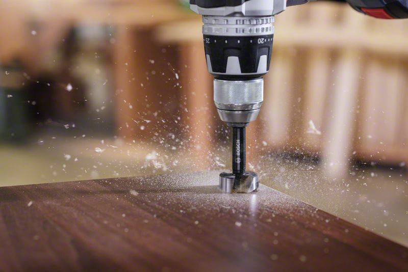 A drill is boring vertically into a wooden board, with wood shavings flying around.
