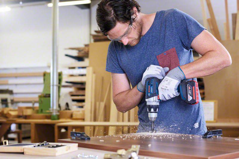 A man in a blue T-shirt is drilling into a wooden board in a workshop. He is wearing safety glasses and gloves.