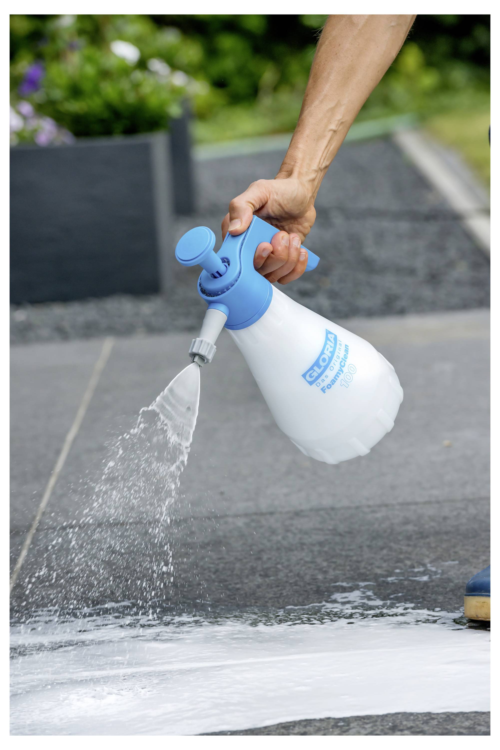 Hand spraying white cleaning foam onto a pavement or concrete surface outdoors with a blue-and-white pressure sprayer.