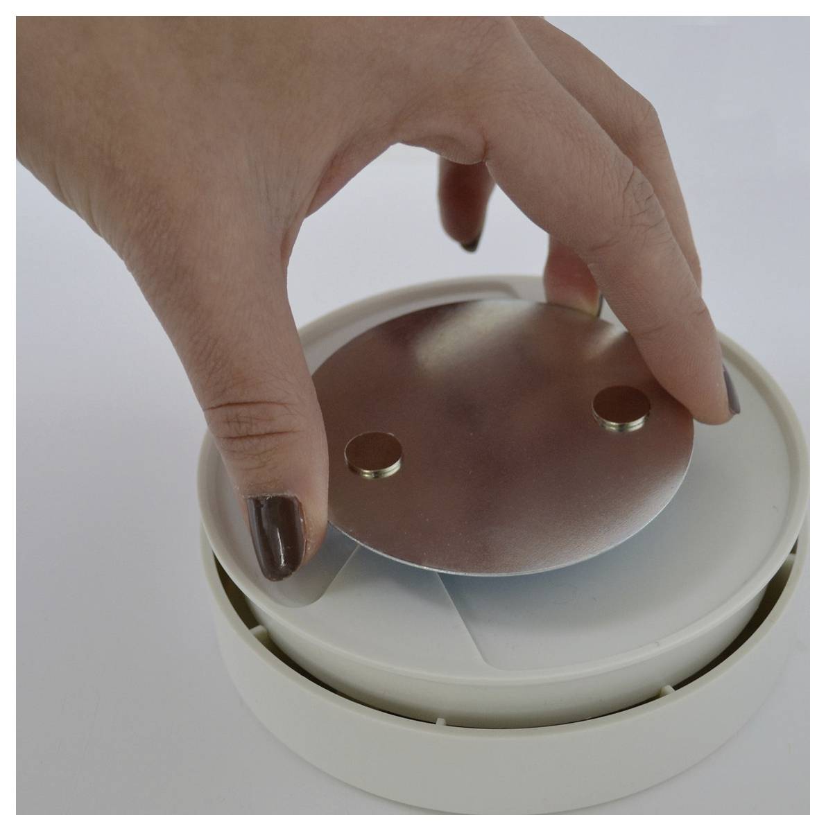 Hand placing a round metallic lid onto a circular white container or device on a plain surface.