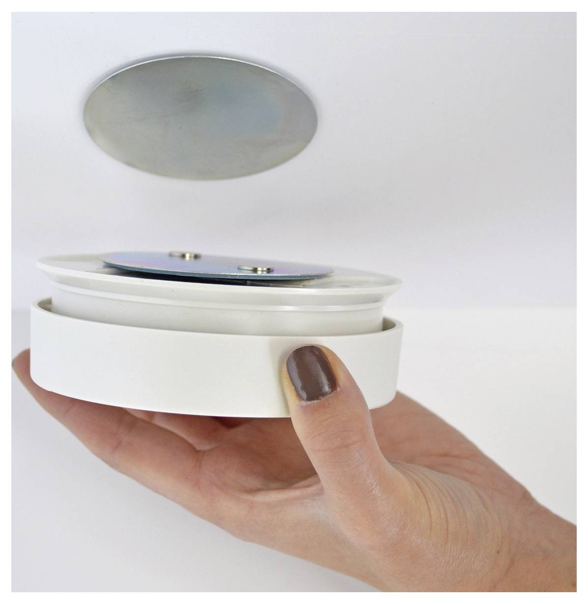 Hand holding a white round device with its lid lifted, revealing a metallic inner disc against a plain background.