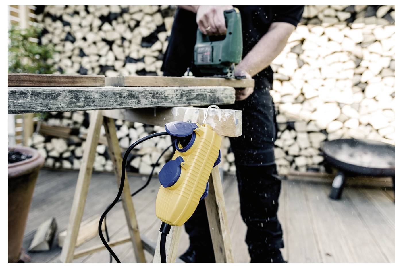 Person using a green power sander on a wooden plank, with a yellow extension cord reel hanging from the workbench in a workshop.<br>