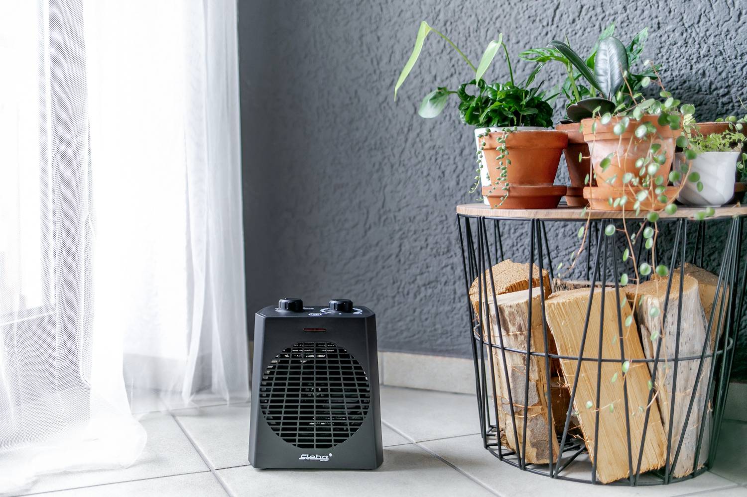 A black fan heater stands on a tiled floor next to a table with potted plants and stacked wood in a living room.
