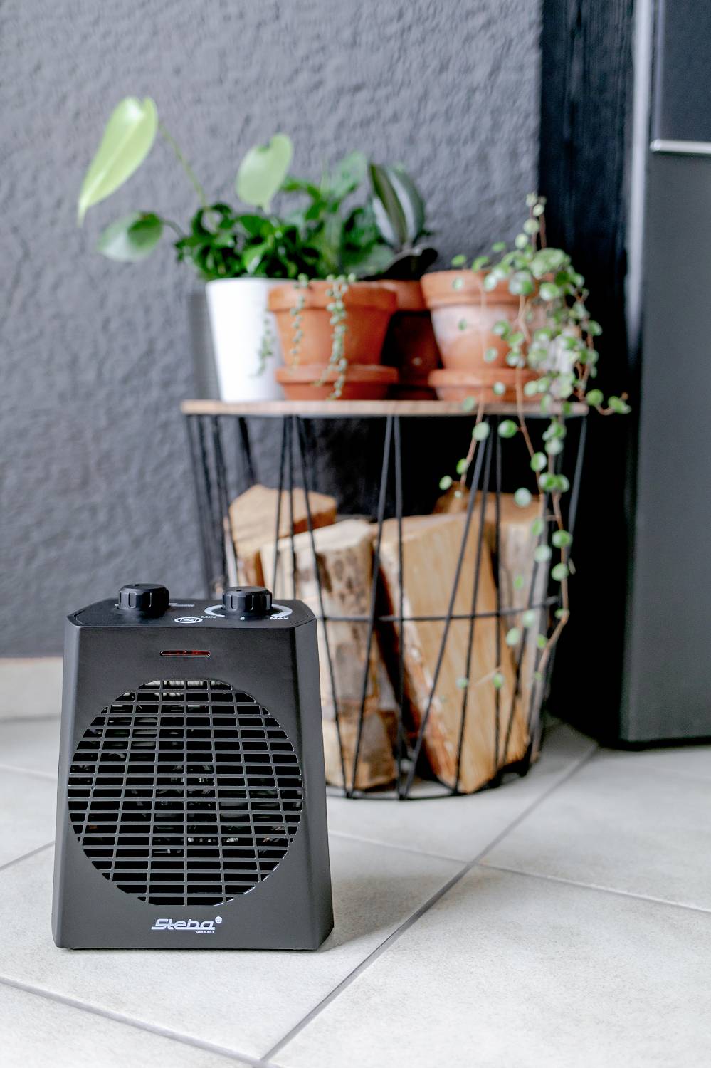 A small black fan heater stands on the floor in front of a side table with potted plants. Background: grey wall.