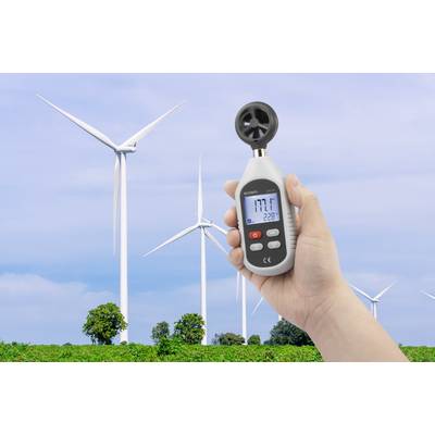 A hand holds an anemometer with a digital display, shown in front of wind turbines in a green field under a blue sky.