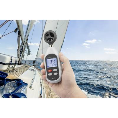 A person is holding an anemometer on a sailing boat; the device is showing '17.7' knots of wind speed under clear skies.