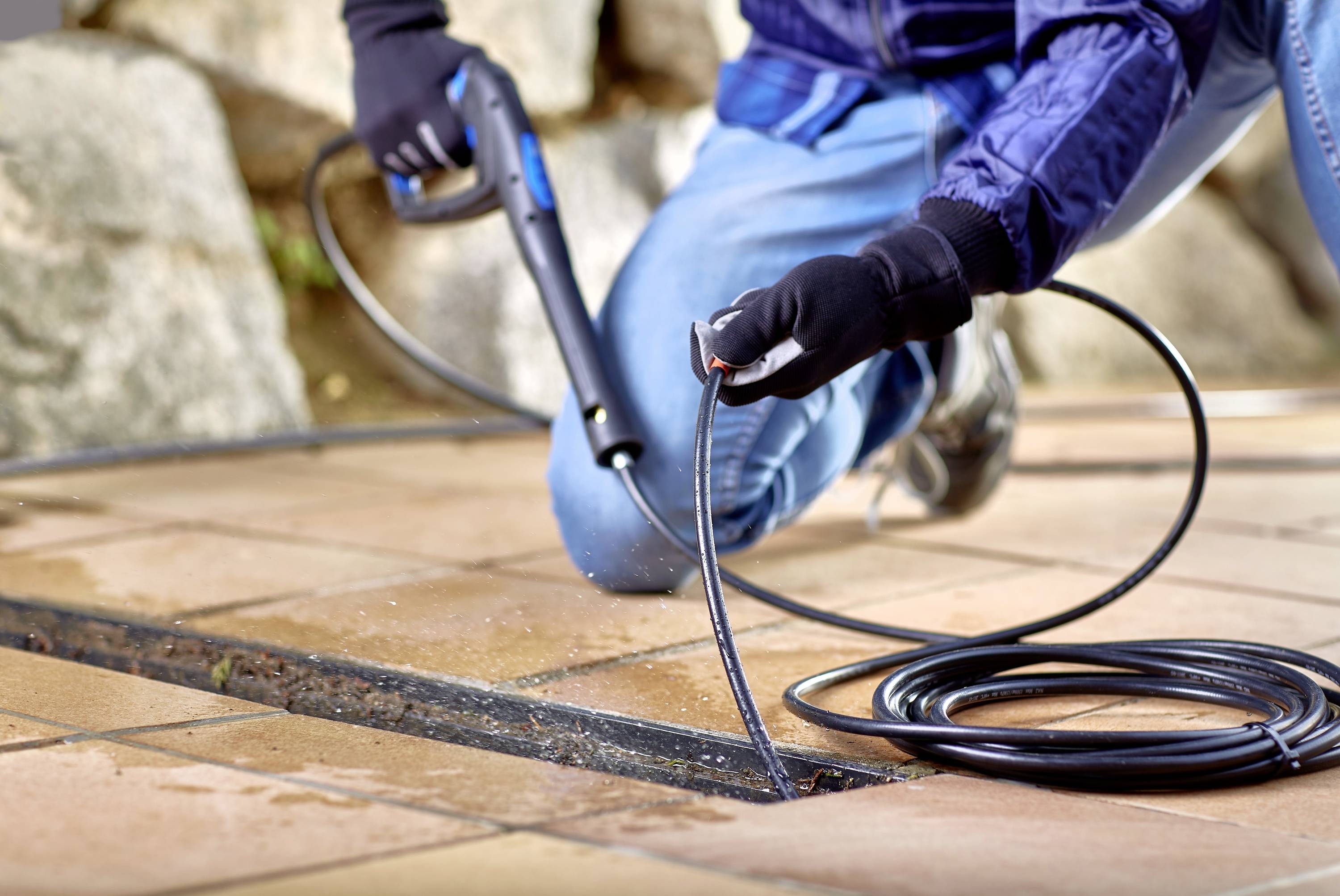 A person is cleaning a tiled outdoor terrace with a pressure washer. Hands, the hose, and tiles are visible.