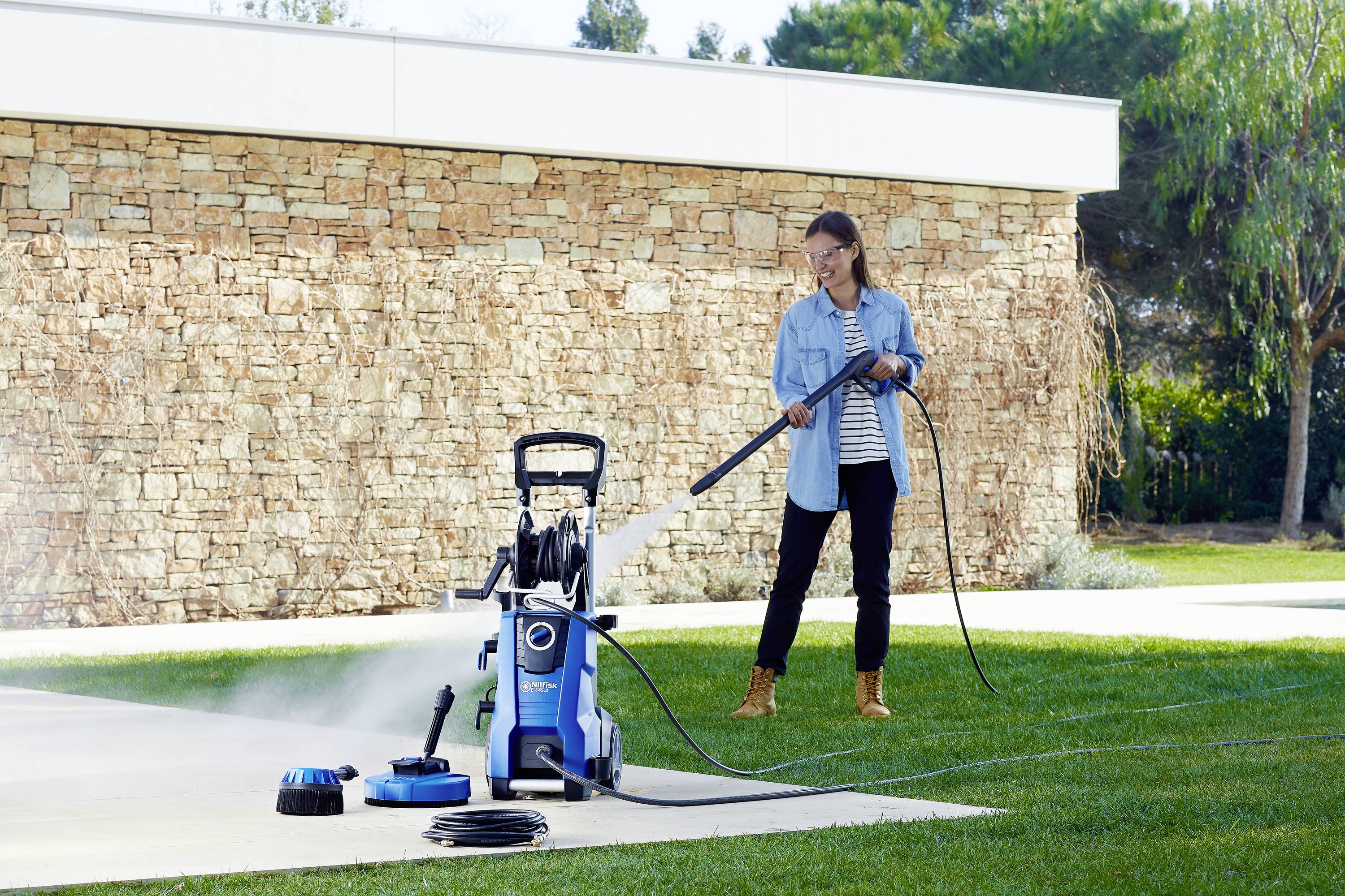 A person is cleaning a patio with a blue pressure washer. A stone wall is visible in the background.