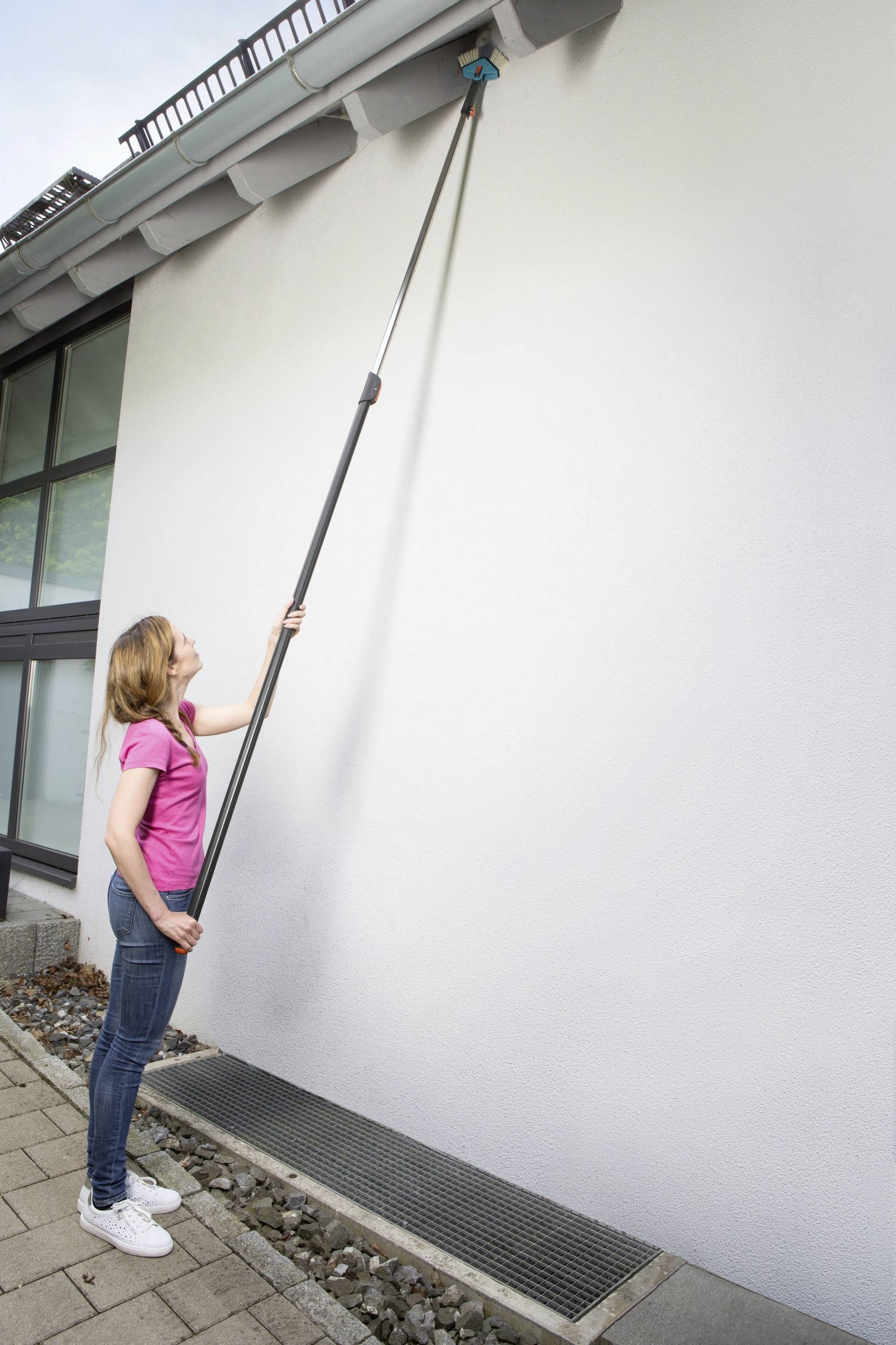 A woman is cleaning the guttering of a house with a long broom. She is wearing a pink T-shirt and blue jeans.