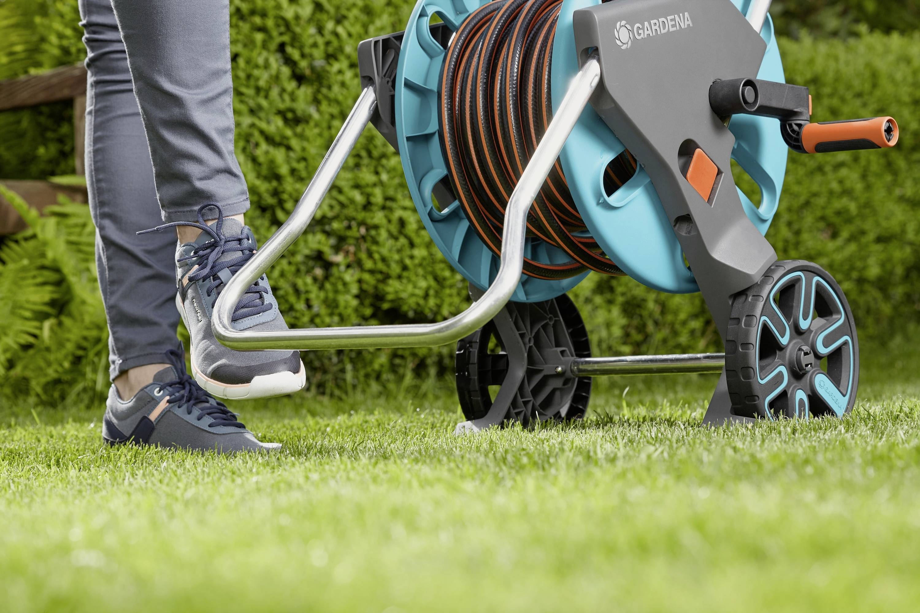 A person steps on the foot lever of a hose reel on a lawn. The trolley has a coiled garden hose.