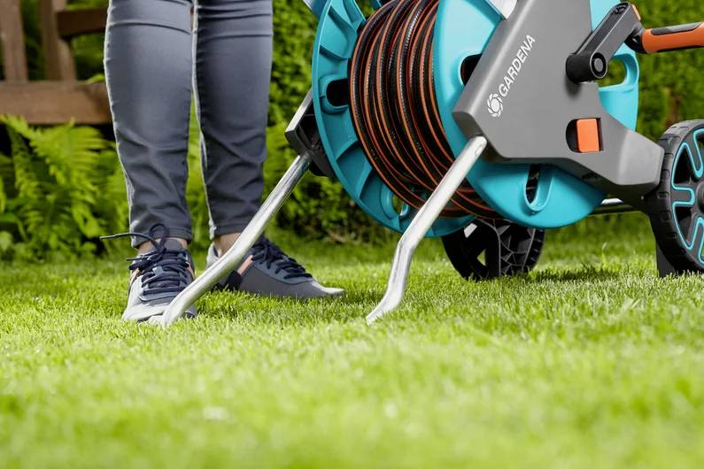 A person uses a Gardena hose reel on a green lawn, wearing jeans and sneakers.