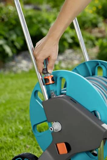 A hand connects a garden hose to a blue and gray hose reel on a grassy lawn, suggesting preparation for watering or gardening tasks.