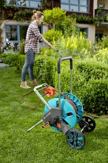 A person in a garden uses a hose reel, watering plants. A hose on a reel with wheels is in the foreground, suggesting easy mobility.