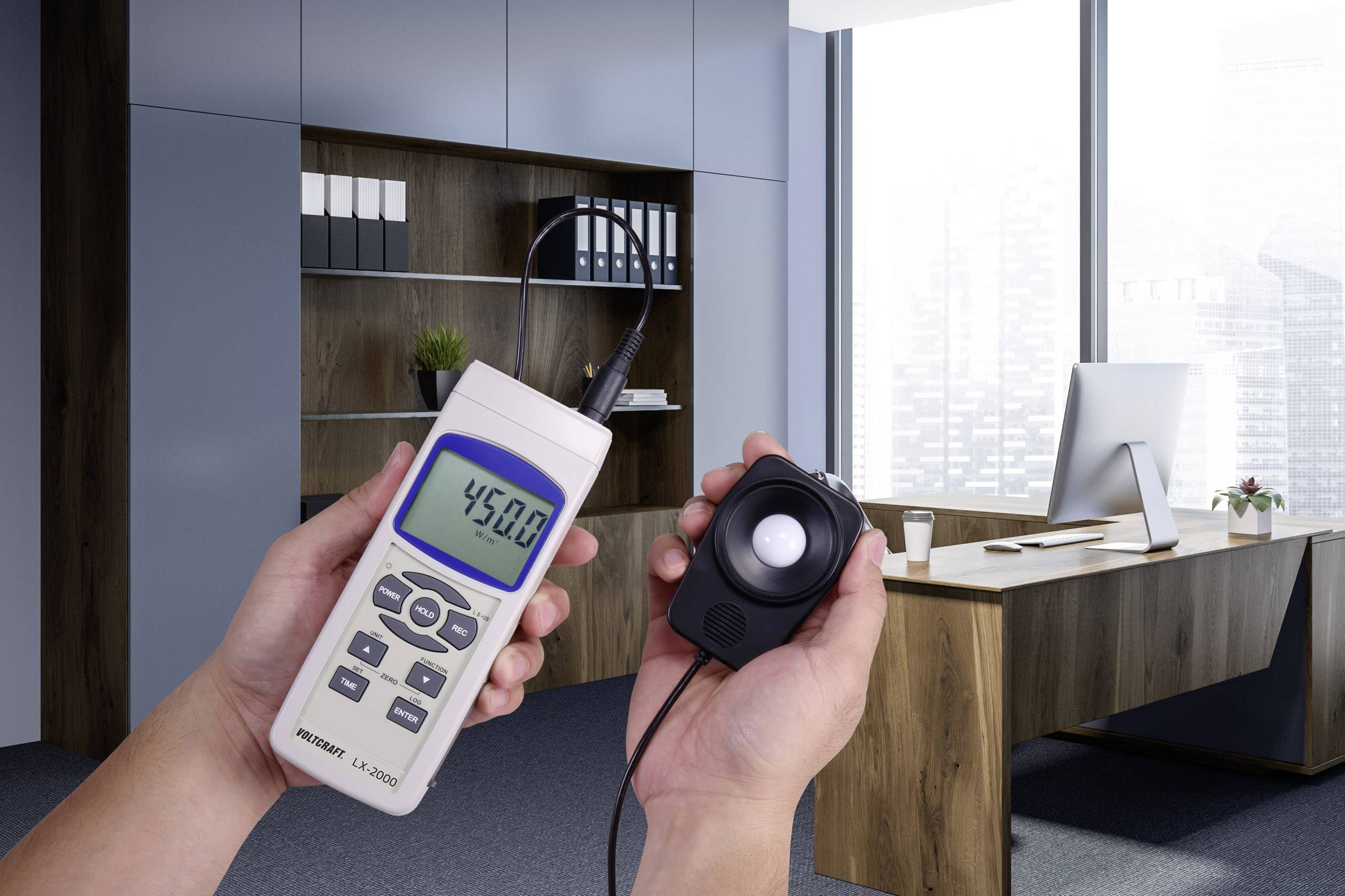 A person is holding a light meter with a display showing '45.0' in a modern office with a desk and computer.