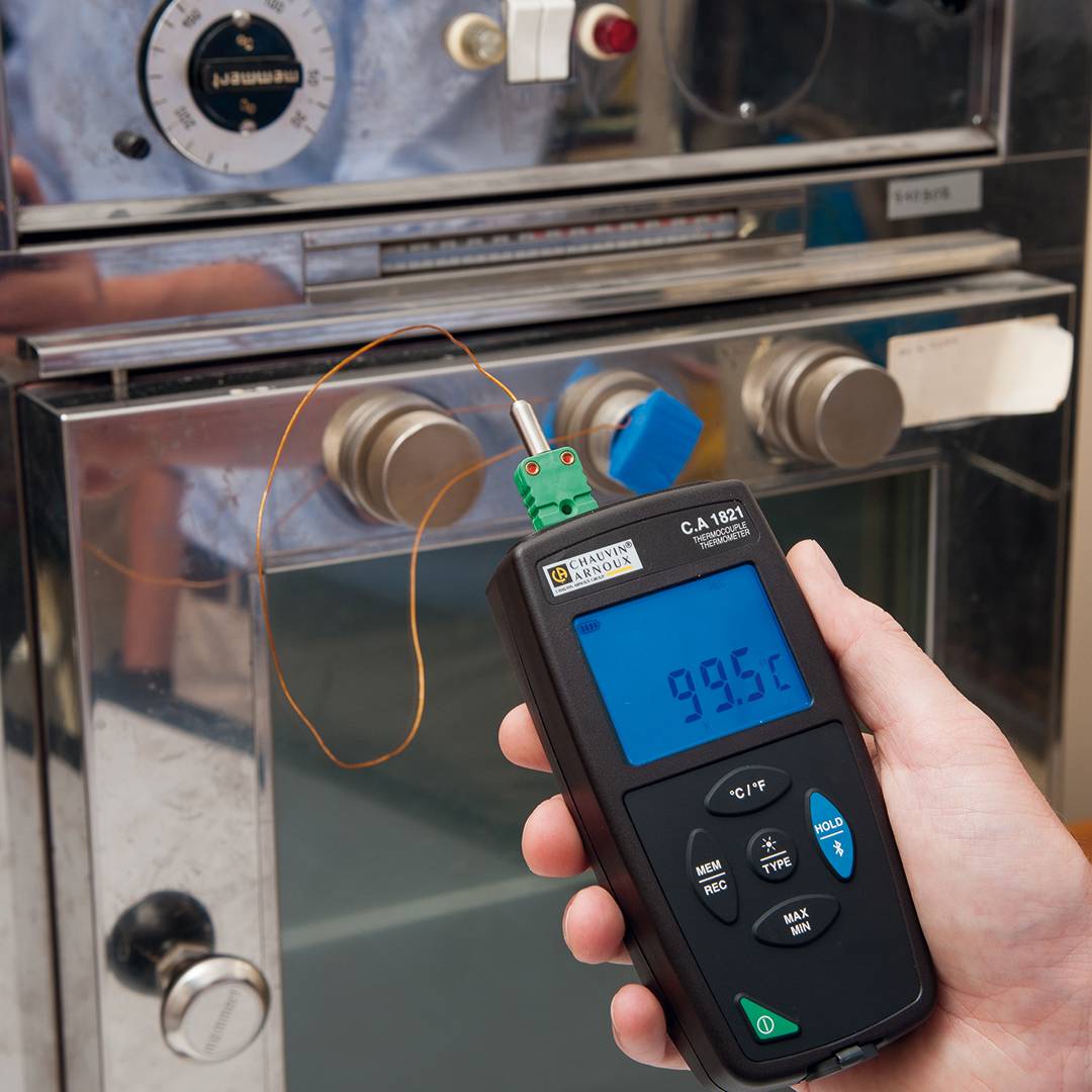 A person is holding a digital thermometer in front of a laboratory oven. The device is displaying a temperature of 99.6 degrees Celsius.