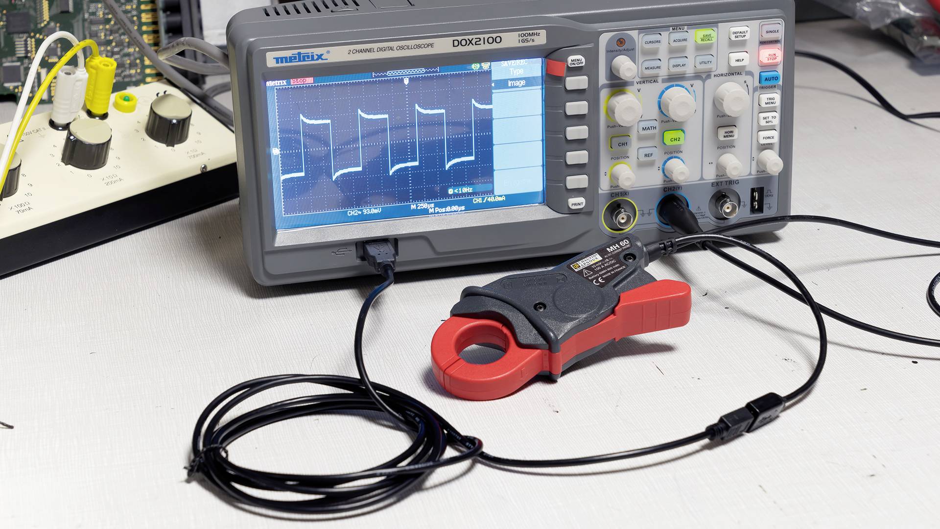 An oscilloscope displays a wave-shaped signal on the screen. Electronic measuring instruments and cables are positioned on the table.