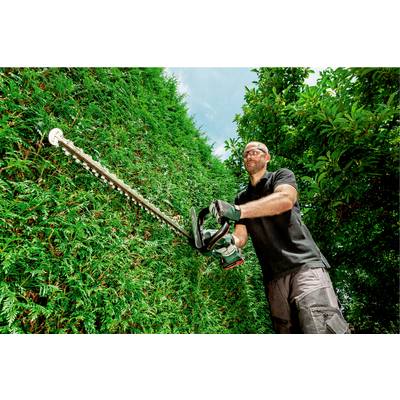 A man is trimming a tall green hedge with a motorised hedge trimmer. He is wearing safety glasses and gloves under a clear sky.