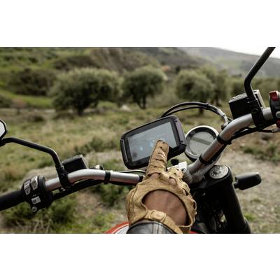 A person wearing leather gloves is tapping on a navigation screen on a motorcycle situated in a rural, wooded setting.