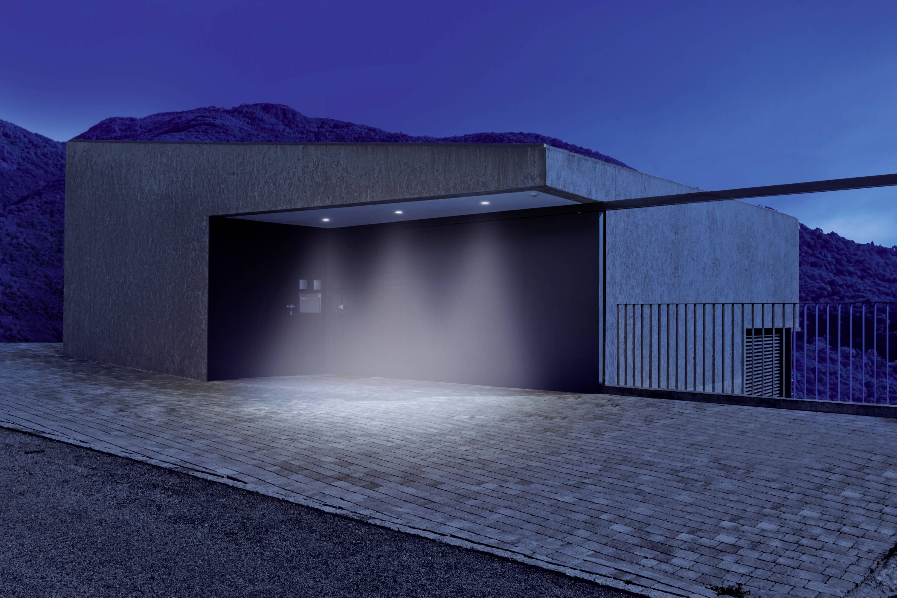 A modern building with an open entrance area, illuminated by exterior lamps, in a mountainous landscape at dusk.