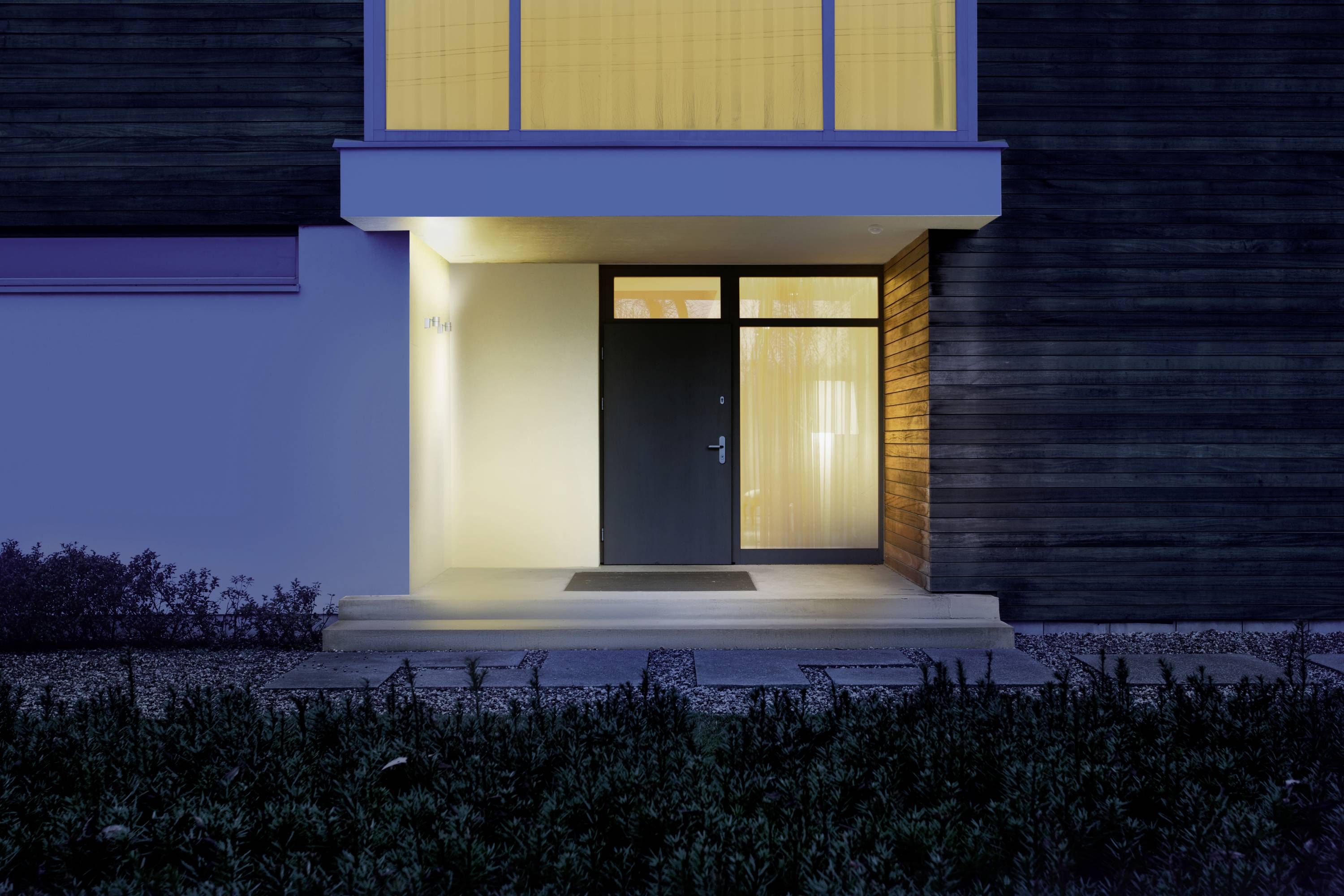 Entrance area of a house at night, illuminated by a yellow outdoor light. Dark façade and large windows above the door.