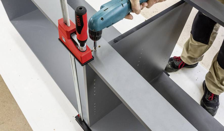 A person is assembling a piece of furniture by securing a grey board with a cordless drill in a red clamp.