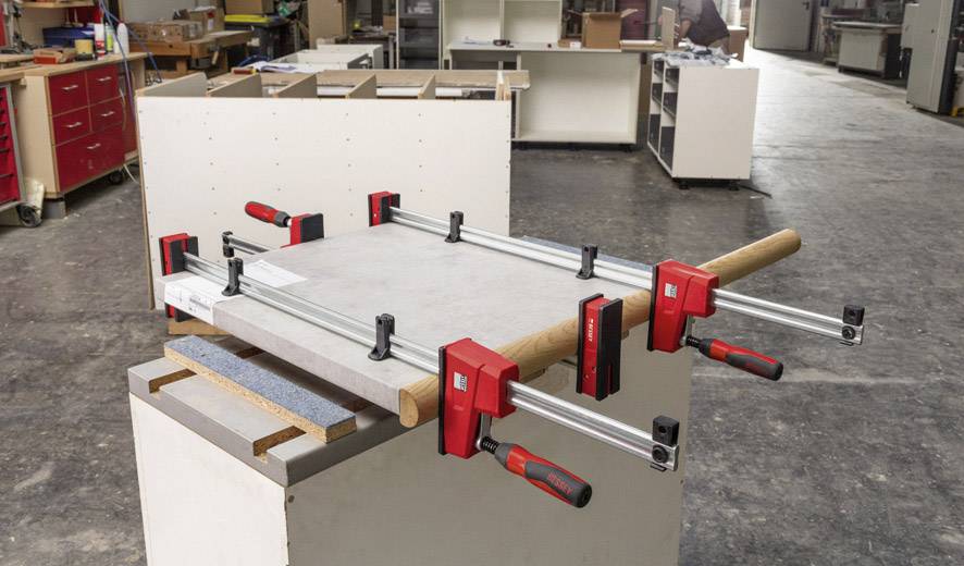 In a workshop, a wooden board is secured to a workbench using several G-clamps. Shelves can be seen in the background.
