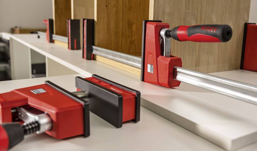 Several red clamps secure wooden boards during furniture assembly on a white table.